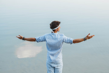 Man handsome dressed in light clothes looks at the water reflex of blue sky flinging his hands to the side. Shoot from the back. Concept of freedom relaxation. Place for text or advertising