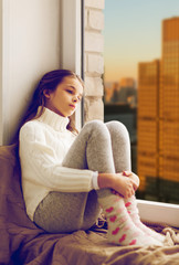 Poster - childhood, sadness and people concept - sad beautiful girl in sweater sitting on sill at home window over city skyscrapers background