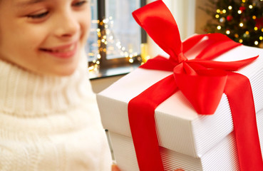 Poster - childhood, holidays and people concept - happy beautiful girl with christmas gift box at home over festive lights background