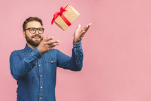 Happy Holiday, My Congredulations! Portrait Of An Attractive Casual Man Giving Present Box And Looking At Camera Isolated Over Pink Background.