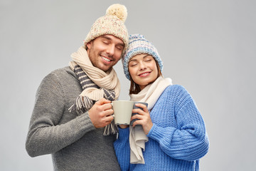 Wall Mural - people, christmas and love concept - happy romantic couple in knitted hats and scarves with mugs over grey background