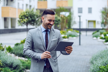 Glamorous smiling caucasian businessman in gray suit using tablet and holding eyeglasses. Public park exterior.