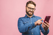 Happy young bearded man in glasses typing sms on pink background.
