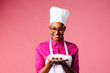 © Carlos David - Portrait of a smiling young female chef holding a plate with three chocolate pralines, isolated on pink studio background