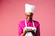 © Carlos David - Portrait of a smiling young female chef looking down at a plate with three chocolate pralines, isolated on pink studio background..