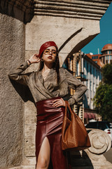 Wall Mural - Outdoor fashion portrait of elegant, luxury woman wearing trendy leather  dark red midi skirt, beret, snakeskin print satin blouse, sunglasses, holding brown handbag, posing in street of European city