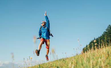 Wall Mural - backpacker traveler emotionally jumping over green grass mountain meadow with backpack with wide ope