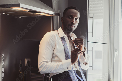 Fotografía Sexy confident male in opened white shirt is putting on his wrist watch while sitting on the kitchen