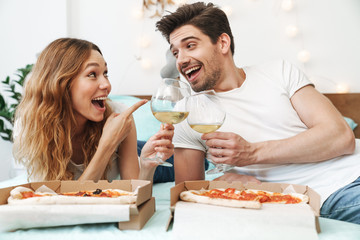 Image of pretty couple drinking wine and eating pizza on bed at home