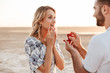 Photo of romantic man making proposal to his amazed woman with ring while walking on sunny beach