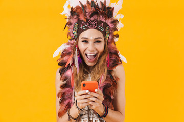 Sticker - Positive young emotional indian woman in carnival suit isolated over yellow wall background using mobile phone.