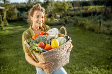 Portrait Of A Beautiful Young Woman With Basket Full Of Freshly Picked Up Vegetables Standing In The Garden On A Sunny Evening