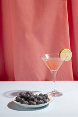 Wall Mural - Martini cocktail glass with lime and blueberries on plate over table near pink curtain