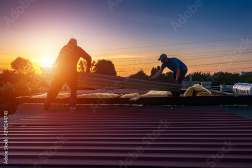 A man holds a board on the roof. Men mount the roof on the building. People are blurred. Focus at the beginning of the picture