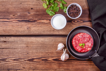 Wall Mural - Raw beef burger meat on a small pan with garlic, salt, pepper and green coriander on a wooden background. Copy space. Top view.