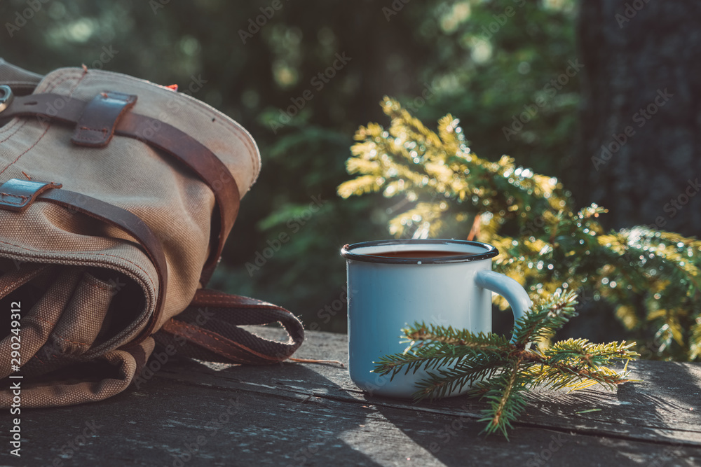 Enameled cup of coffee or tea, backpack of traveller on wooden board in summer forest outdoors. - obrazy, fototapety, plakaty 