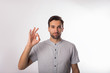 Young handsome bearded business man shows a finger okay and looking in camera while standing isolated in studio