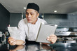 © DragonImages - Asian young chef reading recipe of new dish and using digital tablet while standing in the kitchen