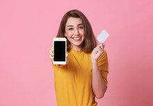 Portrait Of A Happy Young Woman Showing At Blank Screen Mobile Phone And Credit Card Isolated Over Pink Background.