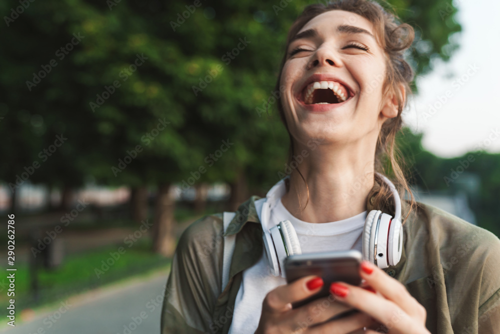 Image of laughing woman holding smartphone and walking in green park - obrazy, fototapety, plakaty 
