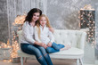 © queen1987 - Two very pretty girls, sisters celebrates Christmas, new year. Girls in the New Year's interior of the room are sitting on a white sofa against the background of garlands