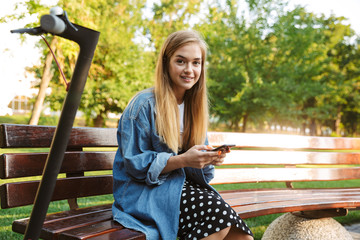 Wall Mural - Cute young teenage girl outside in nature green park with scooter using mobile phone.