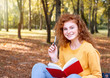 © Nastassia Yakushevic - Smiling happy red hair student girl taking a note outside in autumn park