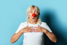 Old Woman With A Red Clown Nose Makes Hand Gestures On A Blue Background. Red Nose Day Concept, Clown, Party