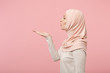 Side view of arabian muslim girl in hijab light clothes posing isolated on pink wall background studio portrait. People religious Islam lifestyle concept. Mock up copy space. Blowing sending air kiss.