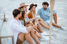 Group Of Young Friends Hanging Out Together With Wine Glasses, Having A Festive Meeting And Enjoying Evening Time On The Sandy Beach