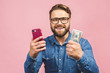 Happy winner! Bearded happy man demonstrating his money prize and using smartphone isolated over pink background.