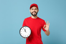 Delivery Man In Red Workwear Hold Clock Isolated On Blue Wall Background Studio Portrait. Professional Male Employee In Cap T-shirt Print Working As Courier Dealer. Service Concept. Mock Up Copy Space