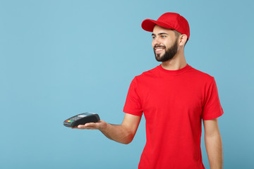 Wall Mural - Delivery man in red workwear hold bank payment terminal to process acquire credit card payments isolated on blue background. Employee in cap t-shirt working courier. Service concept Mock up copy space