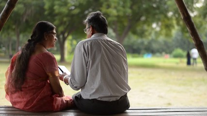 Wall Mural - Senior citizen Indian man reads from mobile and puts hand over shoulder shot from behind on his wife while sitting in a park in Delhi, India