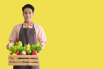 The Handsome young man asian 24s smile wearing brown apron and holding a wood  basket of Non-Toxic fruits and vegetables without chemicals isolated on yellow background. Fruits merchant