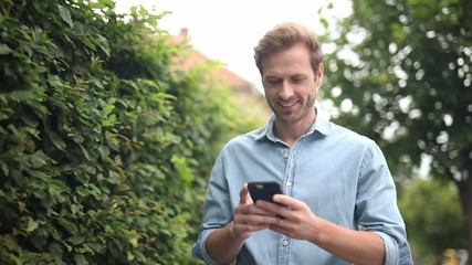 Wall Mural - smiling young casual man reading news on his phone and waves hand as a greeting, in the street