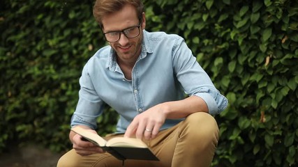 Poster - young casual wearing glasses is reading a book on his knees, on sidewalk