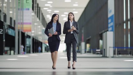 Wall Mural - Full length wide shot of two businesswomen walking down office hall in slow motion and talking. Young woman holding takeaway coffee cup, middle aged colleague carrying laptop computer