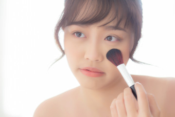 Wall Mural - Beauty portrait young asian woman smile with face looking mirror applying makeup with brush cheek in the bedroom, beautiful of girl holding blusher, skin care and cosmetic concept.