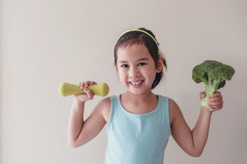 Happy and healthy mixed Asian girl lifting a broccoli and a dumbbell, child wellness,  home exercise, social distance concept