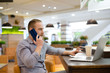 © ruslanshug - young bearded beautiful man calling cellphone business negotiate sitting in cafe with paper cup of coffee and mobile laptop computer