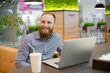 © ruslanshug - happy smiling young bearded caucasian cute man in shirt sitting in cafe with laptop and paper coffee cup during work break