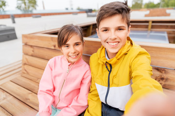 Poster - childhood, technology and people concept - happy children or brother and sister sitting on wooden street bench outdoors taking selfie