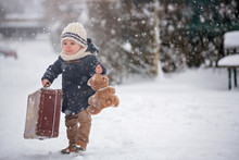 Baby Playing With Teddy In The Snow, Winter Time. Little Toddler Boy In Blue Coat, Holding Suitcase And Teddy Bear, Playing Outdoors In Winter Park
