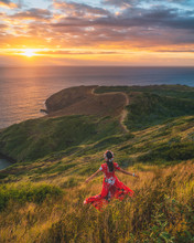 A Woman In Red Flouncing Down A Grassy Hillside Towards The Ocean.