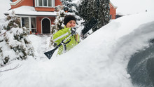 Closeup Photo Of Young Smiling Woman In Green Coat Cleaning A White Snow Covered Car Using A Snow Brush.