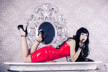 Hot Dominant Brunette Woman In A Red Leather Fetish Dress And Heels Lie On The Table And Look At Camera.