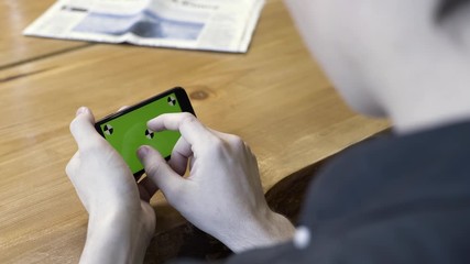 Poster - Smartphone horizontal orientation with a green screen: shot over the shoulder of a young man. Stock Footage. Man sitting at the table, tapping, scrolling, and zooming on the chromakey of the phone.
