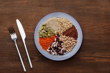 Sticker - top view of fork and knife near striped plate with raw lentil, chickpea, quinoa, oatmeal, beans and pumpkin seeds on wooden surface