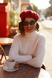 © PinkCoffee Studio - Young beautiful elegant woman in French style cafe wearing in white sweater and red beret sitting in chair and with cup of coffee.